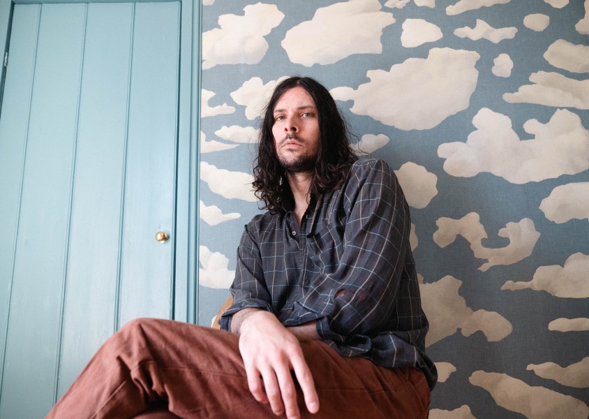 Man sitting against wall with long dark hair, wearing blue check shirt and brown trousersx